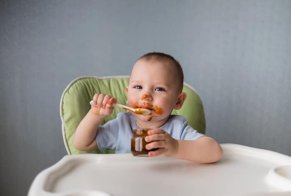 Baby boy eating on his own with a spoon; food all over his face