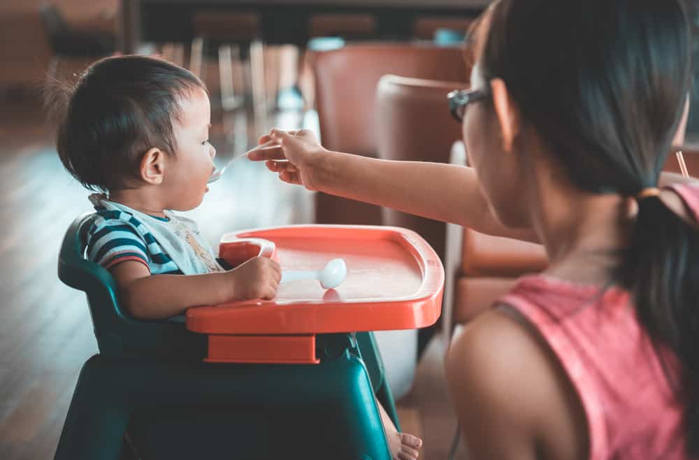 Child sitting in a booster seat being fed by their mother