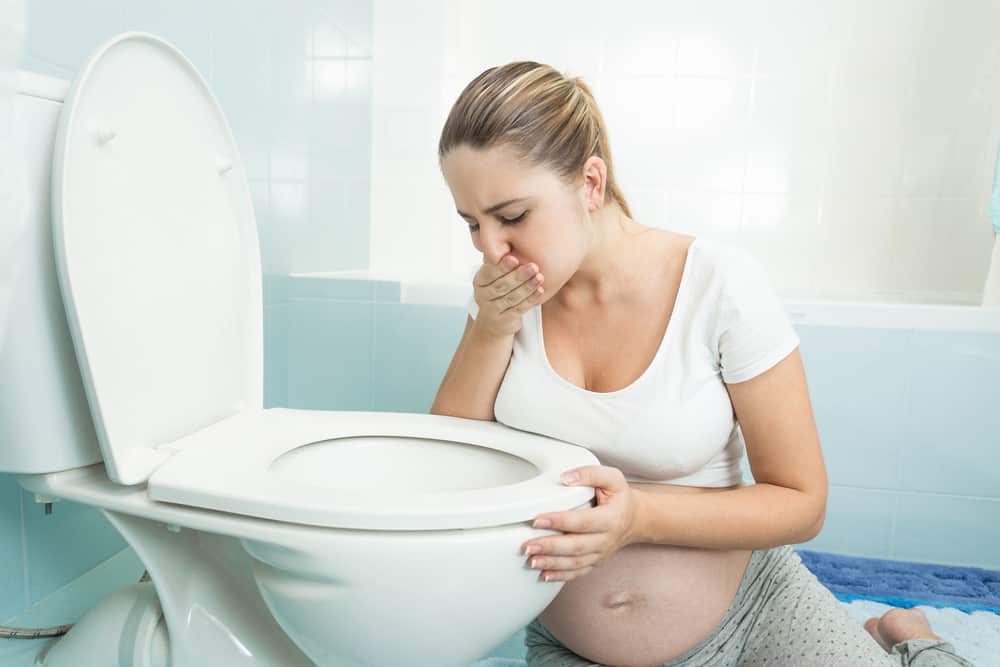 a pregnant woman sits by the toilet with her hand over her mouth feeling sick