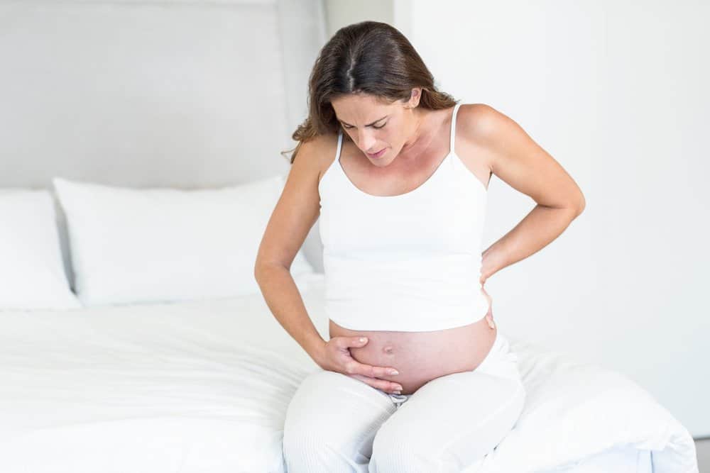 a pregnant woman sits on her bed and holds her stomach in pain