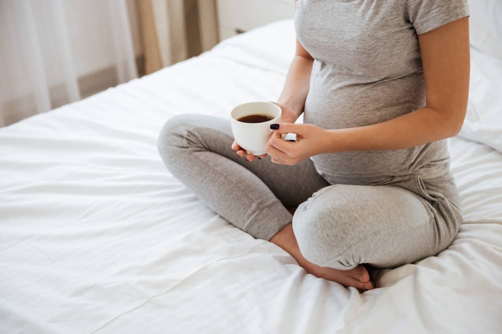 pregnant woman sitting on bed drinking coffee